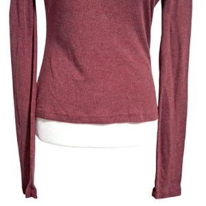 May include: A long-sleeved, burgundy-colored shirt. The shirt has a ribbed texture and a fitted silhouette. The sleeves are long and end with a slight flare at the wrists. The fabric appears to have a subtle sheen.