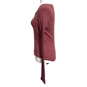 May include: A long-sleeved, burgundy-colored shirt with a V-neck and a decorative trim along the neckline. The shirt has a slim fit and a long, flowing tail.
