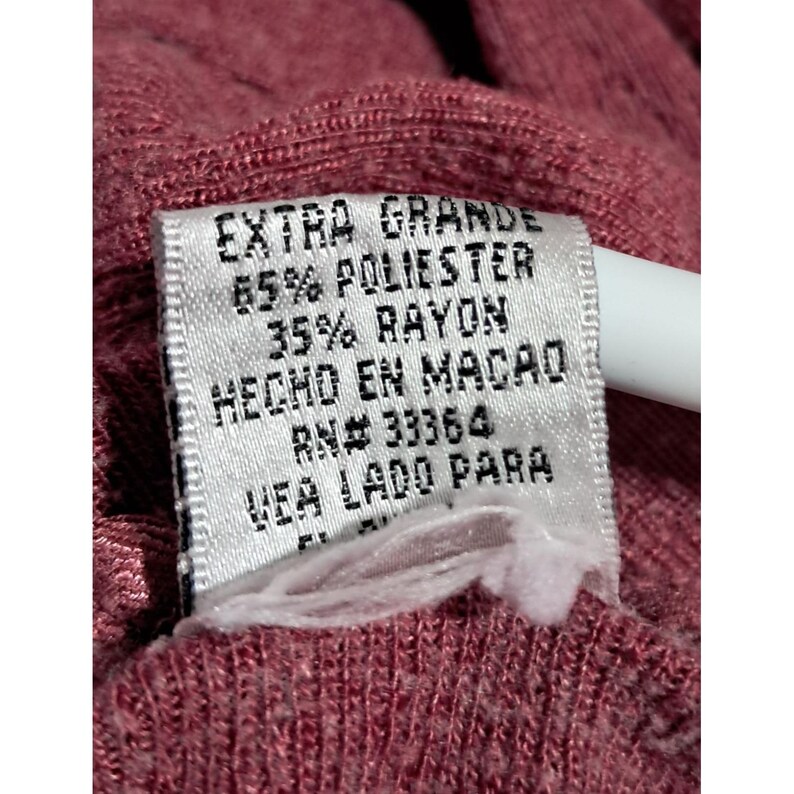 May include: Close-up of a tag with the words "EXTRA GRANDE" and fabric composition details: 65% POLIESTER, 35% RAYON. The tag also includes "HECHO EN MACAO" and "RN# 33364". The tag is attached to a maroon fabric.