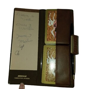 May include: A brown leather card holder with a pen and two decks of playing cards. The holder has a flap with a metal clasp and a pocket for a pen. The cards are in a plastic sleeve. The holder has the text "BOEING FABRICATION DIVISION" printed on the front.