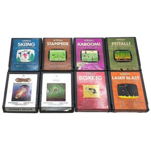 May include: A collection of eight rectangular Activision video game cartridges for the Atari 2600 console. The titles include Skiing, Stampede, Kaboom!, Pitfall!, Journey Escape, Bugs, Boxing, and Laser Blast, each featuring unique cover art.