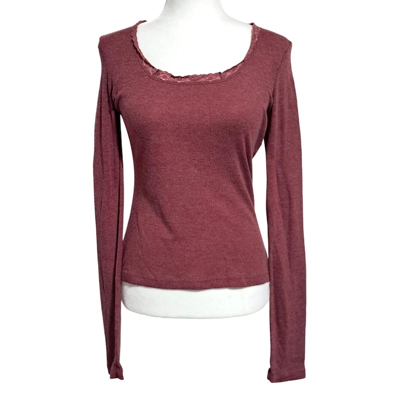 May include: A long-sleeved burgundy shirt with a scoop neck and a delicate lace trim. The shirt is displayed on a mannequin, showcasing its form-fitting design and soft fabric. A casual top for everyday wear.
