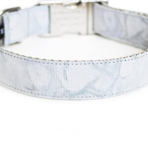 May include: A light gray dog collar with a marbled pattern. The collar has a silver-colored metal buckle and hardware. A small black tag with white text is sewn onto the collar.