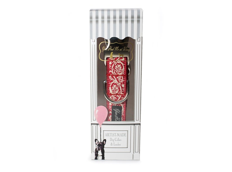 May include: A red and white patterned dog collar and leash set displayed in a decorative box. The collar features a floral design. The box has a striped top and a drawing of a dog with a balloon. Text on the box reads "Artist-Made Dog Collar & Leash."