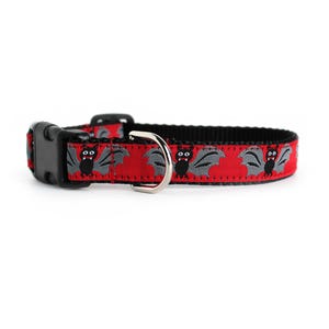Puede incluir: Un collar para mascota de color rojo con un patrón repetitivo de murciélagos grises con cuerpos negros y colmillos blancos. El collar incluye una hebilla de plástico negro de liberación lateral y una anilla en D de metal plateado.