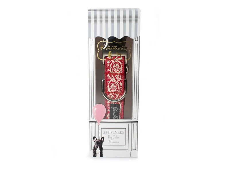 May include: A red and white patterned dog collar and leash set displayed in a decorative box. The collar features a floral design. The box has a striped top and a graphic of a dog holding a balloon. Text on the box reads "Artist-Made Dog Collar & Leash."