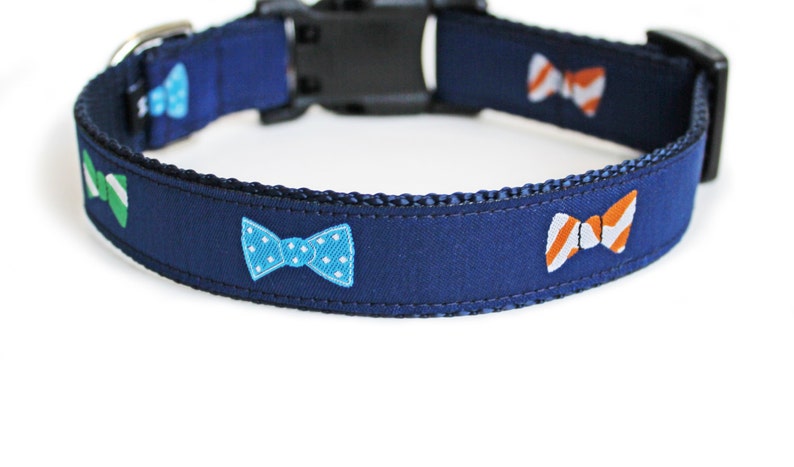 May include: A navy blue dog collar with a repeating pattern of colorful bow ties. The bow ties are green, blue with white polka dots, and orange and white striped.