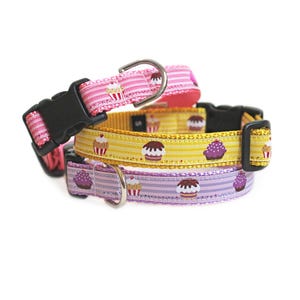 Puede incluir: Tres collares para mascotas con estampados de cupcakes en rosa, amarillo y morado. Cada collar incluye una hebilla de plástico negra, un ajustador y una anilla en D de metal plateado para enganchar la correa. Los collares están apilados.