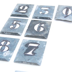 Puede incluir: Un conjunto de diez plantillas de números de metal, cada una con un número diferente del 0 al 9. Las plantillas son de color plateado y tienen un aspecto ligeramente desgastado, vintage.