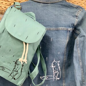 Op de afbeelding: Een lichtblauwe rugzak met een kattenillustratie en een spijkerjasje met een bijpassend kattenontwerp. De rugzak heeft een klepsluiting en een trekkoord, en de jas heeft een klassieke kraag.