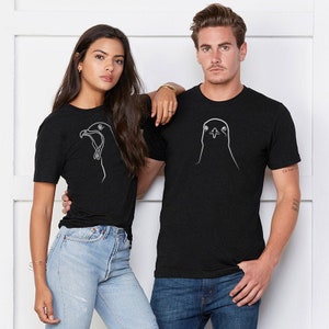 Pode incluir: Um casal vestindo camisetas pretas com um desenho de linha branca de uma cabeça de pássaro em cada camiseta. A mulher está vestindo jeans azul e o homem está vestindo jeans azul.