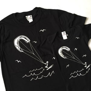 Pode incluir: Duas camisetas pretas com um gráfico branco de uma pessoa fazendo kitesurf. A camiseta maior tem o texto "Kite Clothing" na etiqueta.