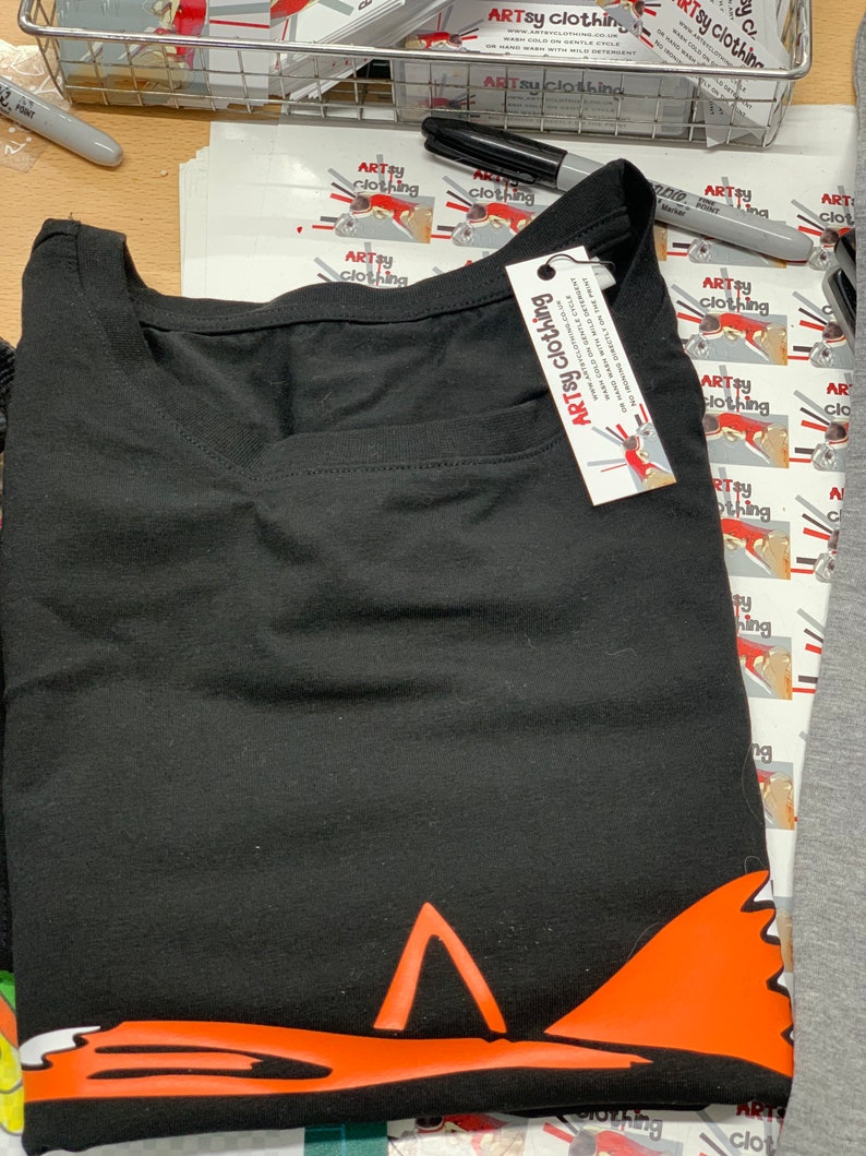 K&ouml;nnte beinhalten: Schwarzes T-Shirt mit einem roten Fuchs-Motiv am unteren Rand. Das Shirt hat ein Etikett mit der Aufschrift "ARTsy clothing".