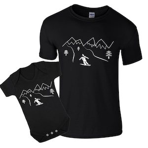 K&ouml;nnte beinhalten: Ein schwarzes T-Shirt und ein schwarzer Baby-Strampler mit einer wei&szlig;en Umrandung eines Skifahrers, der einen Berg mit B&auml;umen und Bergen im Hintergrund hinunterf&auml;hrt.