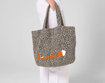 Könnte beinhalten: Eine Tragetasche mit grau-schwarzem Leopardenmuster und einer stilisierten orange-weißen Fuchsgrafik, die horizontal im unteren Bereich platziert ist. Die Tasche verfügt über zwei passende Tragegriffe im Leopardenmuster und eine weite Öffnung.