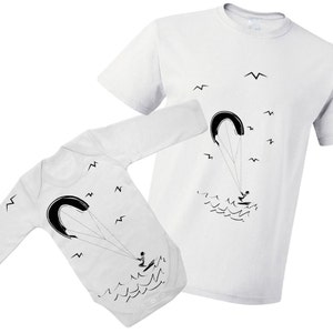 Pode incluir: Body branco de manga comprida e camiseta com uma ilustração em preto e branco de uma pessoa a fazer kitesurf. A ilustração inclui um papagaio, uma pessoa numa prancha de surf e ondas. Também há pequenos pássaros negros a voar no céu.