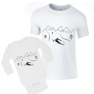 Pode incluir: Um macacão branco de manga comprida para bebê e uma camiseta branca de manga curta com um desenho de linha preta de um esquiador descendo uma montanha com árvores e montanhas ao fundo.