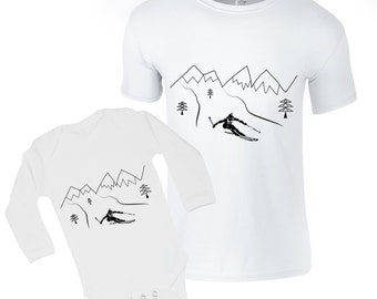 May include: A white long-sleeved baby onesie and a white short-sleeved t-shirt with a black line drawing of a skier going down a mountain with trees and mountains in the background.