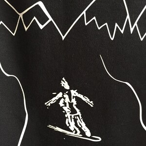 K&ouml;nnte beinhalten: Schwarz-wei&szlig;-Grafikdruck eines Skifahrers auf schwarzem Hintergrund. Der Skifahrer tr&auml;gt eine Jacke und eine Hose. Der Skifahrer f&auml;hrt einen Berg hinunter. Der Hintergrund ist ein stilisiertes Gebirge.