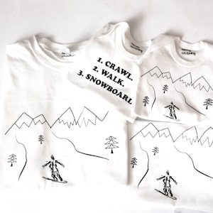Pode incluir: Três camisetas brancas com desenhos pretos de montanhas e esquiadores. A camiseta maior tem o texto "1. CRAWL. 2. WALK. 3. SNOWBOARD."