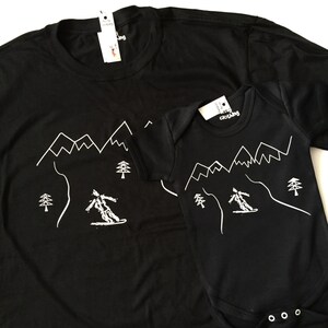 K&ouml;nnte beinhalten: Schwarzes T-Shirt und Strampler mit einer wei&szlig;en Umrandung eines Skifahrers, der einen Berg mit B&auml;umen hinunterf&auml;hrt. Der Text "merry clothing" befindet sich auf dem Etikett jedes Kleidungsst&uuml;cks.
