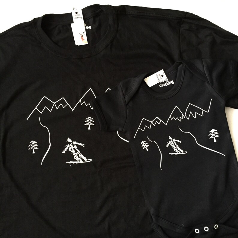 K&ouml;nnte beinhalten: Schwarzes T-Shirt und Strampler mit einer wei&szlig;en Umrandung eines Skifahrers, der einen Berg mit B&auml;umen hinunterf&auml;hrt. Der Text "merry clothing" befindet sich auf dem Etikett jedes Kleidungsst&uuml;cks.