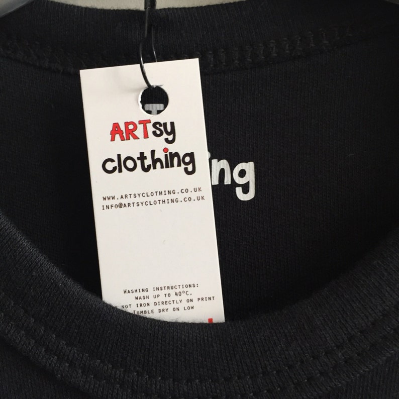 K&ouml;nnte beinhalten: Ein wei&szlig;es Etikett mit schwarzem Text, der "ARTSY clothing" und "WWW.ARTSYCLOTHING.CO.UK" und "INFO@ARTSYCLOTHING.CO.UK" lautet. Das Etikett ist an einem schwarzen T-Shirt befestigt, auf dem das Wort "ing" aufgedruckt ist.