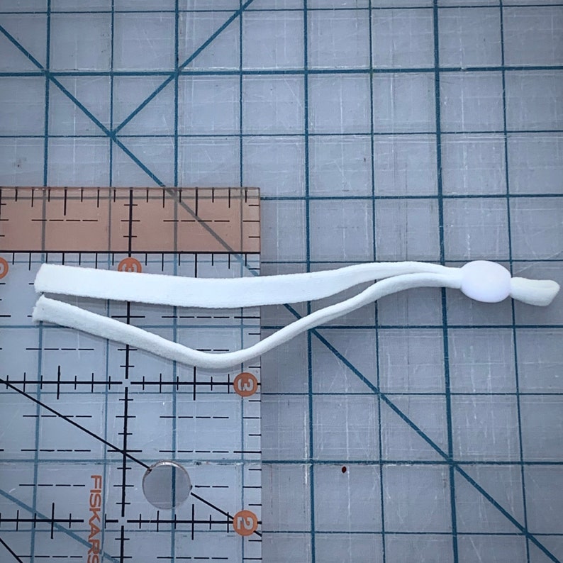 May include: White elastic cord with a white plastic toggle. The cord is about 10 inches long and is measured against a ruler.