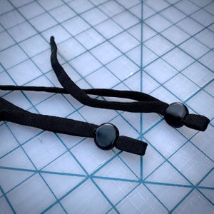 May include: Two black elastic cords with black plastic adjustable toggles. The cords are lying on a white grid surface.