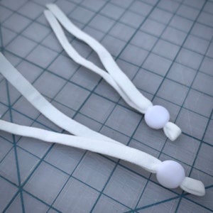 May include: Two white elastic straps with white plastic toggles at the ends. The straps are lying on a gray grid surface.