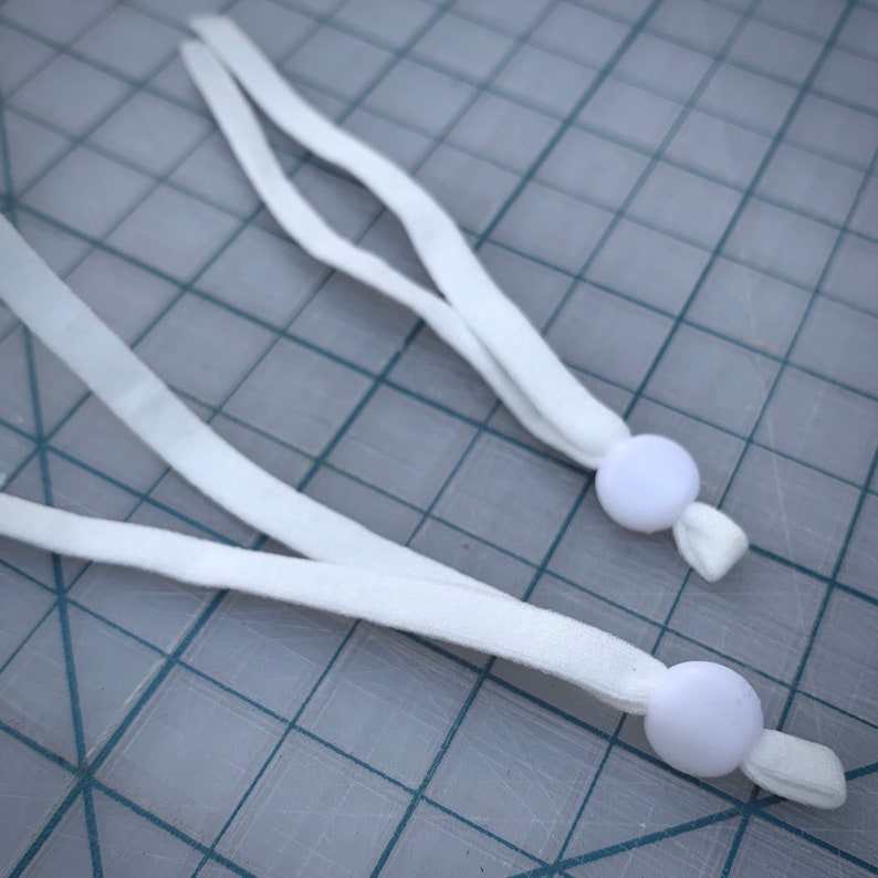 May include: Two white elastic straps with white plastic toggles at the ends. The straps are lying on a gray grid surface.