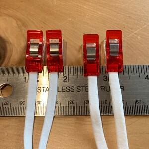 May include: Four red plastic clips holding two white elastic bands in place. The elastic bands are measured against a ruler, showing the length of each band is approximately 3 inches.