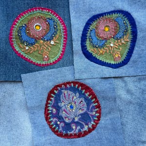 May include: Three denim fabric squares featuring circular embroidered patches. Two patches have green centers with pink and blue floral embroidery and yellow gems. One patch has a dark blue center with purple floral embroidery and a blue gem.