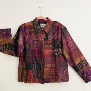 May include: A patchwork jacket in shades of burgundy, brown, and black, with embroidered geometric designs. The jacket has a collared neckline, long sleeves, and button closures. The brand "Chicos" is visible on the label.