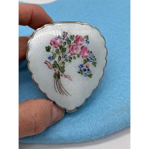 May include: A silver-toned compact with a floral design. The compact is heart-shaped with a scalloped edge. The lid features a bouquet of pink roses, blue forget-me-nots, and green leaves tied with a pink ribbon.