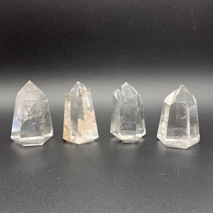 May include: Four clear quartz crystal points with faceted sides and pointed tops. The crystals are arranged in a row against a dark gray background. Each crystal has a unique internal pattern and slight variations in clarity.