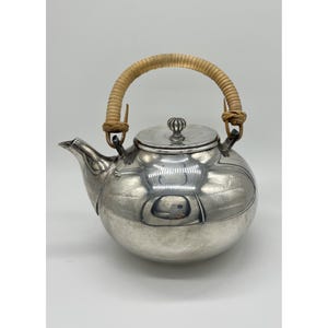 May include: A silver teapot with a woven handle. The teapot has a round body, a spout, and a lid with a small knob. The handle is made of a light brown woven material. The teapot is reflective and appears to be made of metal.
