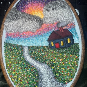 May include: Hand-stitched embroidery art in an oval wooden hoop. The scene depicts a colorful sunset, fluffy clouds, a small house, and a path through a field of flowers. The house has a red door and yellow lit windows. The sky transitions from purple to orange.
