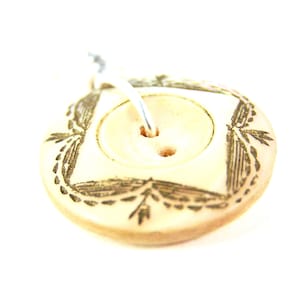 May include: A cream-colored button with a square design and two holes. The button has a decorative, dark-colored leaf pattern around the square. A white thread is looped through the button.