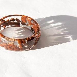 May include: A clear resin bangle bracelet with brown coffee beans embedded inside. The bracelet is on a white surface.