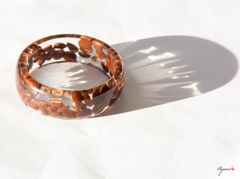 May include: A clear resin bangle bracelet with brown coffee beans embedded inside. The bracelet is on a white surface.