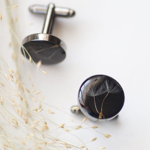 May include: A pair of silver cufflinks with black resin insets. One cufflink features a dandelion seed head, the other has a geometric design.