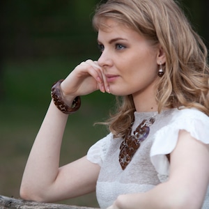 May include: A woman with long blonde hair wearing a white dress with a brown and white necklace. She is wearing a brown bracelet on her wrist and has a thoughtful expression on her face.