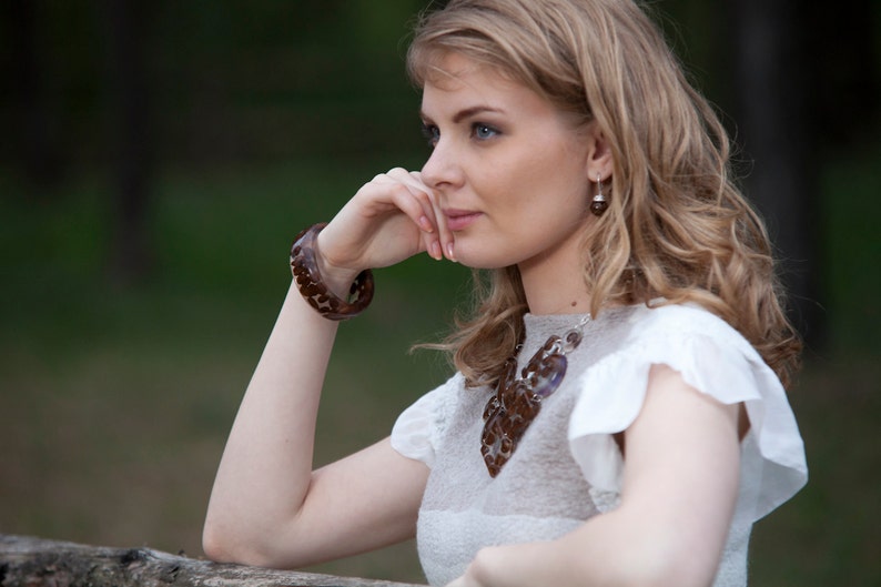 May include: A woman with long blonde hair wearing a white dress with a brown and white necklace. She is wearing a brown bracelet on her wrist and has a thoughtful expression on her face.