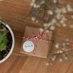 Op de afbeelding: Een klein bruin cadeaudoosje gebonden met rood en wit touw. Een witte ronde sticker met het woord "Agnera" in cursief staat op de doos.