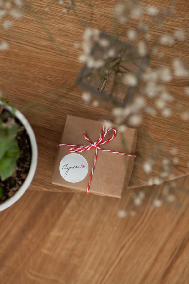 Op de afbeelding: Een klein bruin cadeaudoosje gebonden met rood en wit touw. Een witte ronde sticker met het woord "Agnera" in cursief staat op de doos.