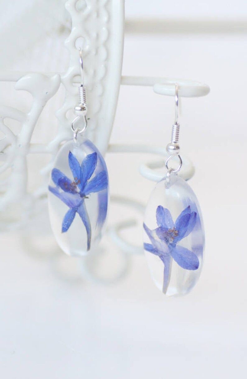 Op de afbeelding: Een paar zilveren oorhangers met heldere hars ovalen met geperste blauwe bloemen.