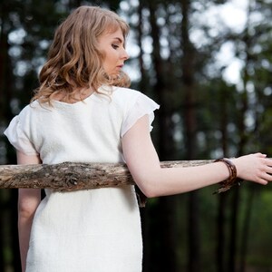 May include: A woman in a white dress with a brown bracelet stands in a forest with a wooden fence behind her.