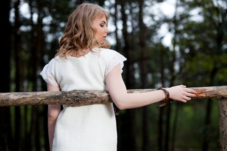 May include: A woman in a white dress with a brown bracelet stands in a forest with a wooden fence behind her.