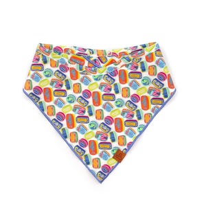 Può includere: Una bandana bianca con un motivo colorato di forme rettangolari e circolari nei toni dell'arancione, blu, verde e rosa. La bandana ha un bordo blu e un'etichetta marrone.